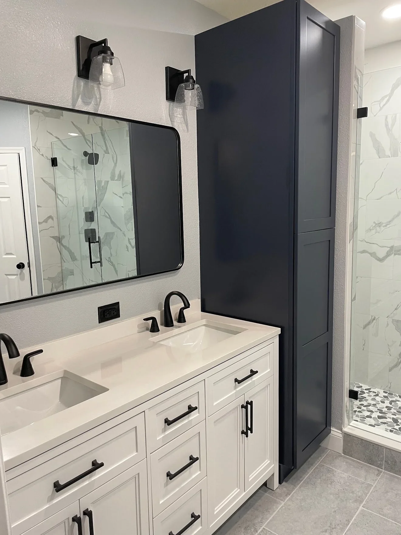 White vanity with quartz countertop, black-framed mirror, marble tile walls, and glass shower enclosure