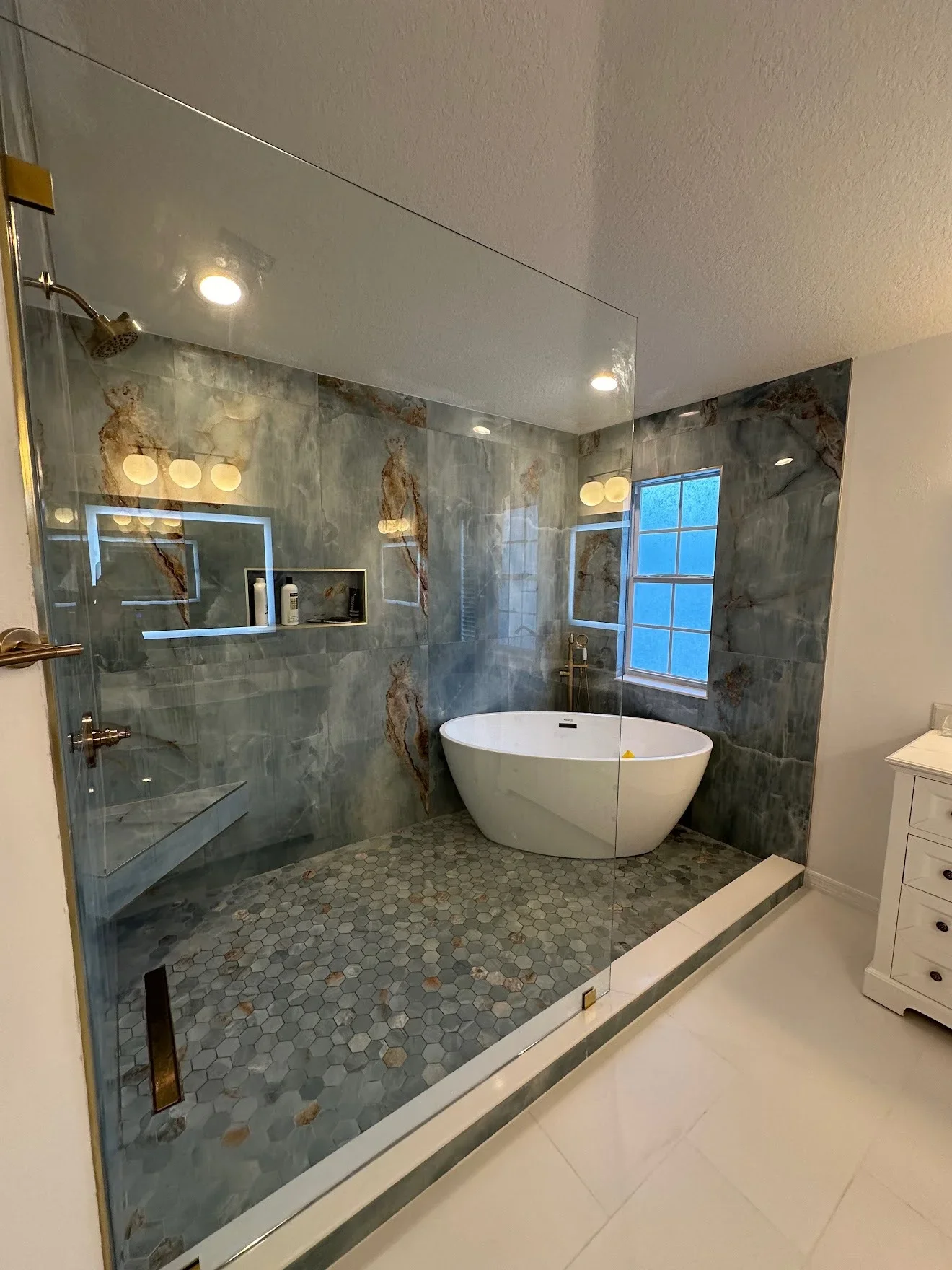 Frameless glass shower enclosure with slate tile walls, freestanding tub, hexagonal floor tile, and recessed lighting