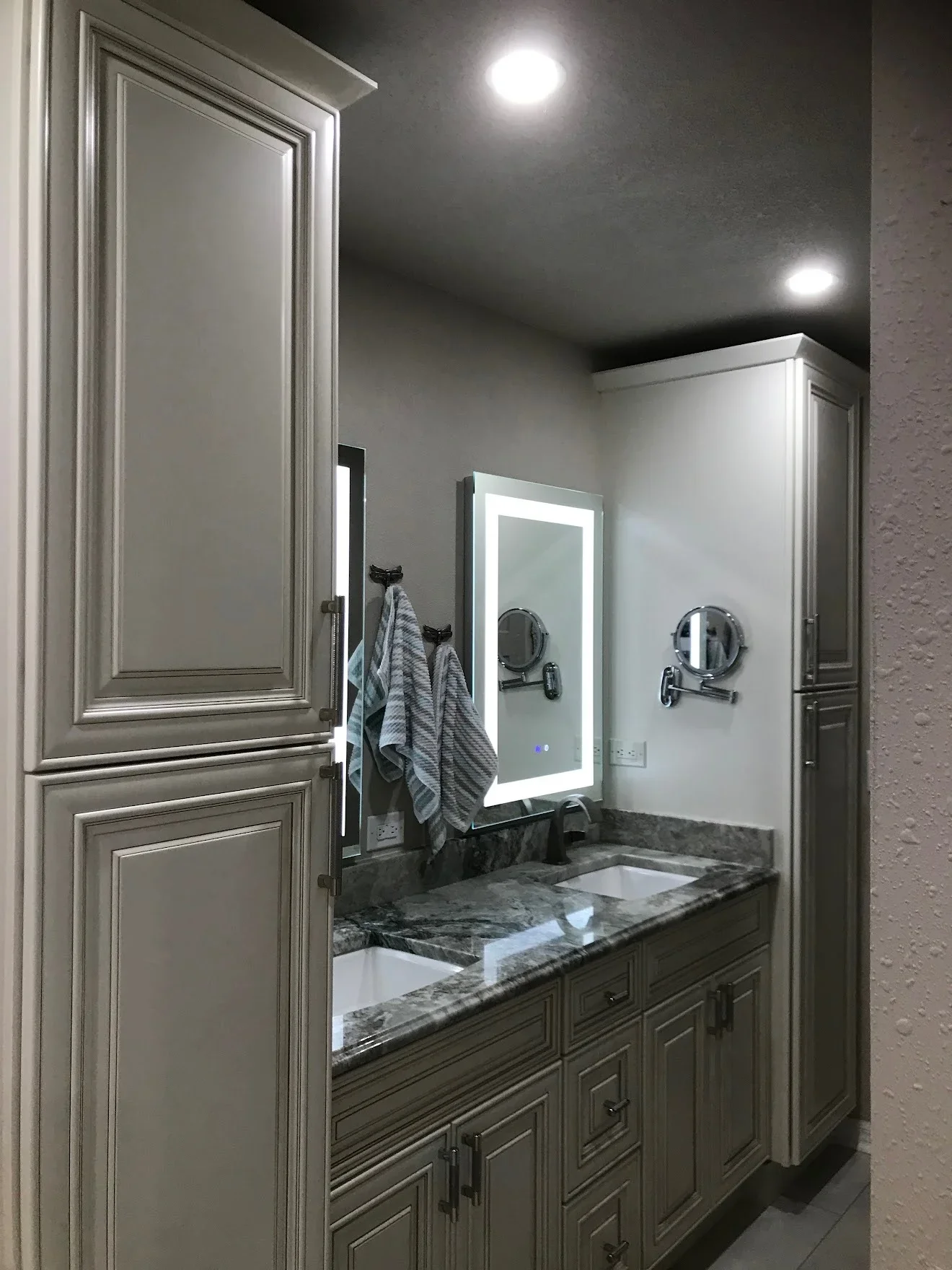 Dual-sink bathroom vanity with granite countertop, custom cabinetry, and backlit mirror