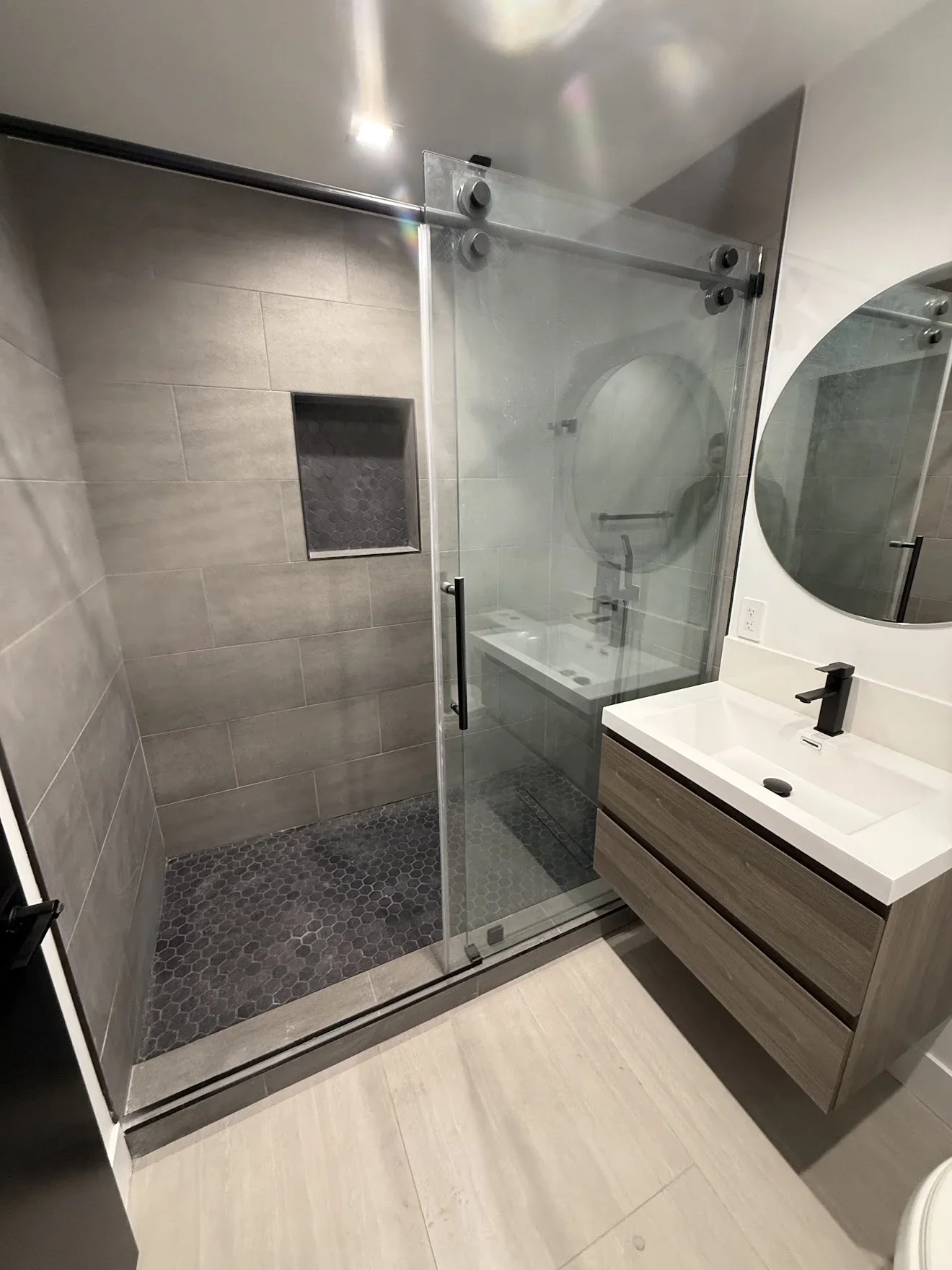 Modern bathroom with gray tile walls, frameless glass shower, and floating wood vanity with white sink