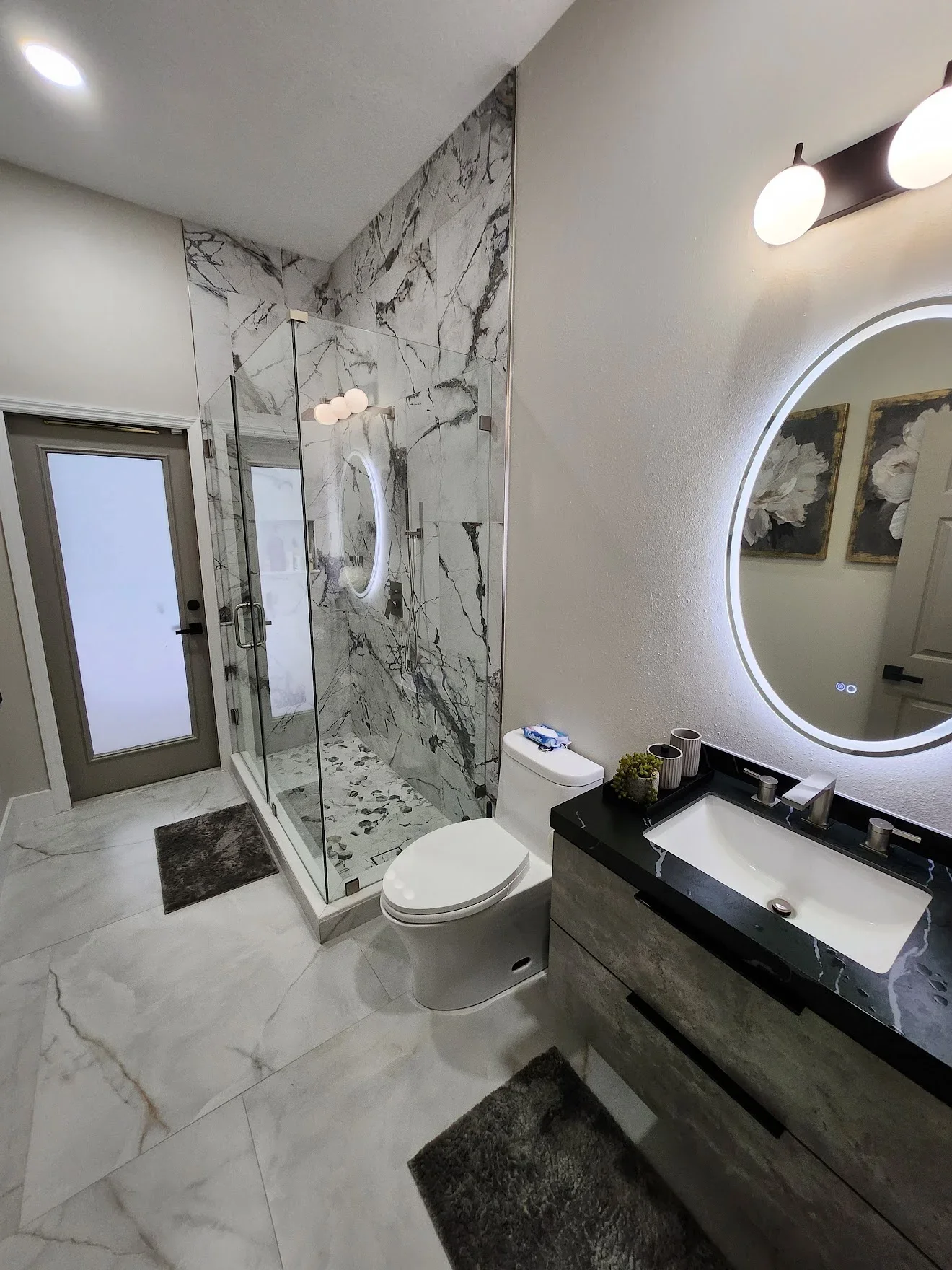 Marble tile shower with frameless glass doors, marble flooring, black vanity with white sink, and LED mirror