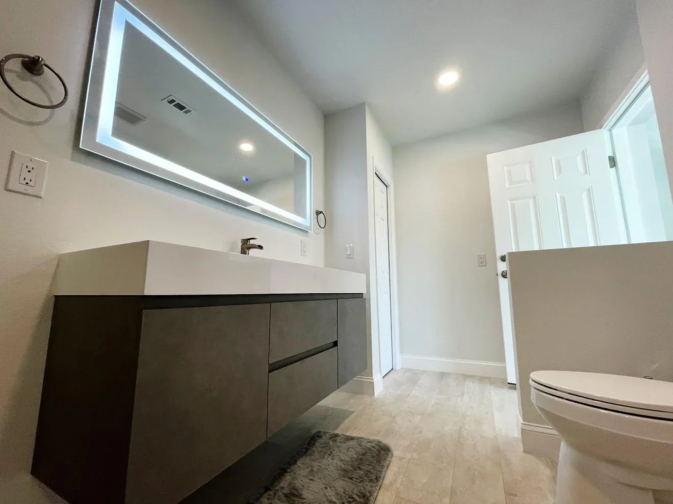 Modern bathroom with dark vanity, white quartz countertop, LED-backlit mirror, and light wood-look flooring