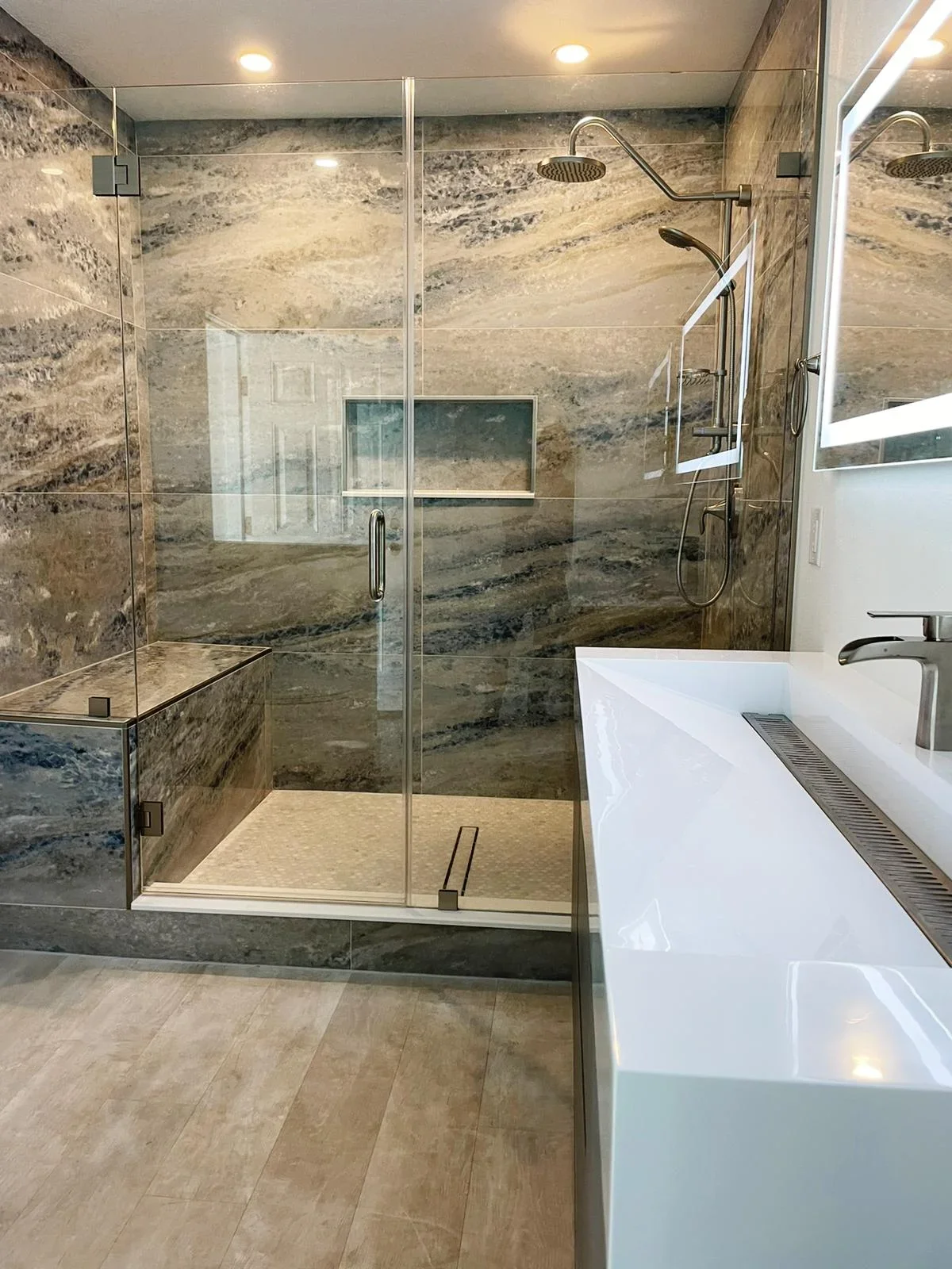 Spacious walk-in shower with travertine tile walls, frameless glass doors, dual showerheads, and white vanity